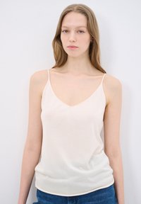 Light beige camisole top with thin straps, v-neckline, soft texture, and a fitted silhouette. Paired with blue jeans.