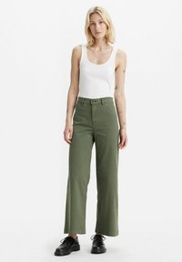 Grüne weite Baumwollhosen mit hoher Taille, einem Knopfverschluss und einer glatten Textur, kombiniert mit einem figurbetonten weißen Tanktop.