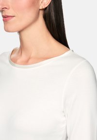 Weißes Langarmoberteil mit weitem Ausschnitt, glatter Textur und genähten Akzenten am Ausschnitt und den Armausschnitten, vor einem neutralen Hintergrund präsentiert.