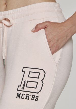 Hand in pocket of light pink sweatpants with elastic waist and black "Bench B MCR'89" logo printed on thigh.