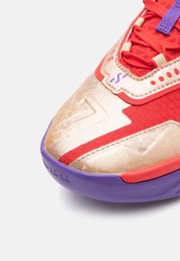 Buty sportowe z teksturowaną, złotą i czerwoną cholewką, z purpurowymi akcentami, siateczkowym językiem i skomplikowanymi detalami przeszycia.