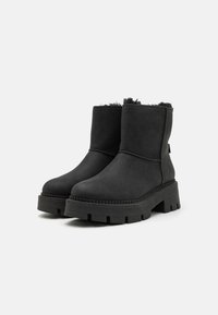 Botas negras hasta el tobillo hechas de material suave, con una suela de goma gruesa con detalles estriados y un borde de piel sintética en la parte superior.