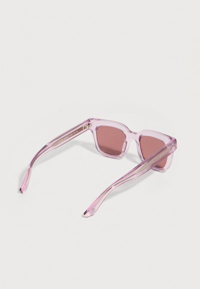 Ροζ διαφανή τετράγωνα γυαλιά ηλίου με ροζ φακούς, ανοιχτά σε λευκή επιφάνεια.