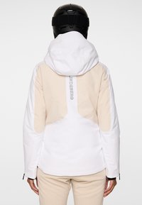 Osoba ubrana w beżową i białą kurtkę z kapturem oraz czarny kask, widoczna z tyłu, z widocznym napisem marki na kurtce.