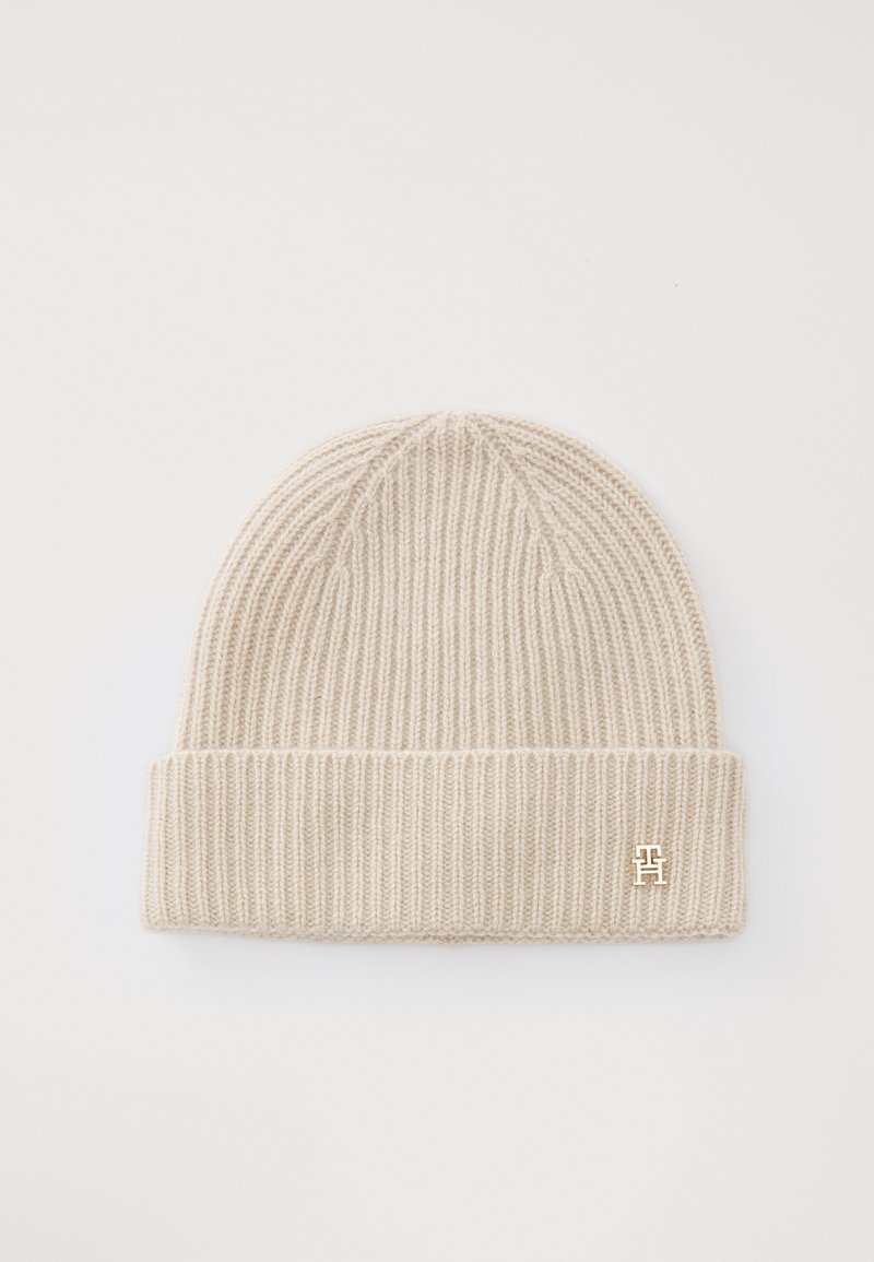 Beige strikket lue laget av ribbet stoff, med en brettet kant og en liten gulllogo på siden. Myk tekstur.