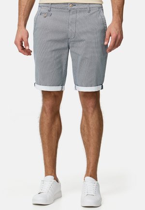 Homme portant un short gris à motifs avec les revers roulés et des baskets blanches, debout devant un fond uni.