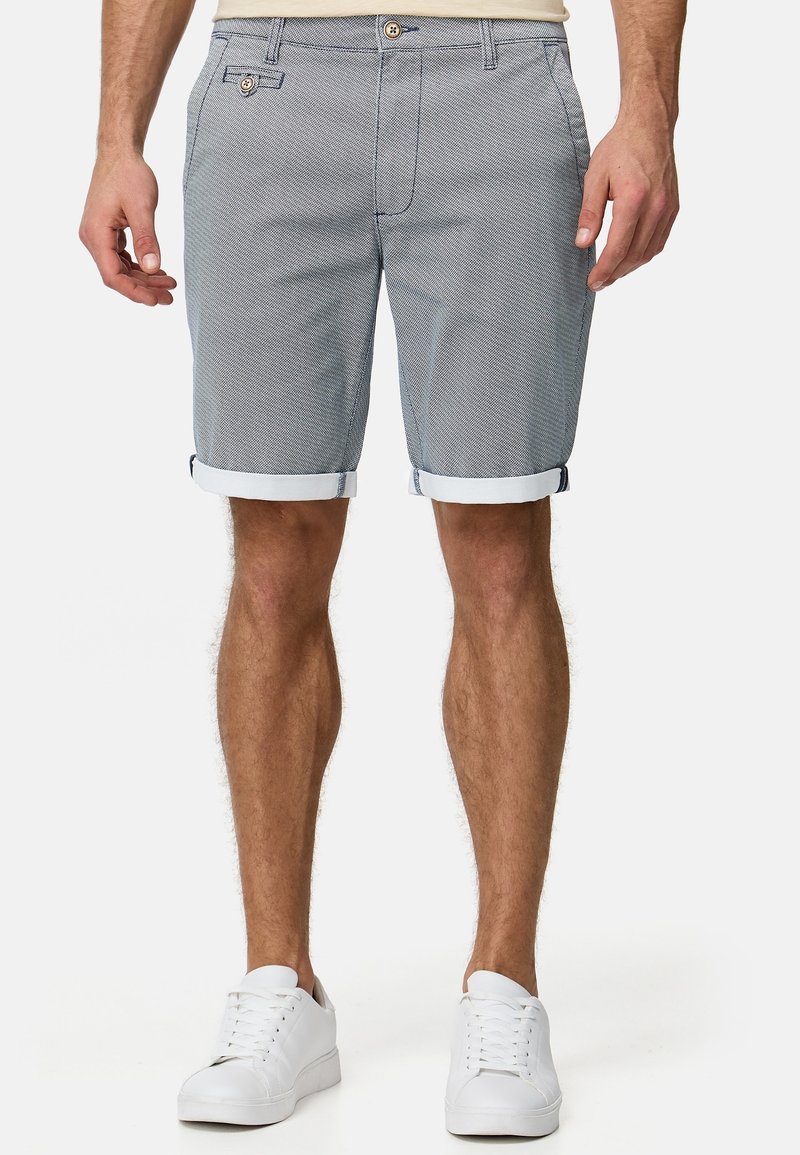 Homme portant un short gris à motifs avec les revers roulés et des baskets blanches, debout devant un fond uni.