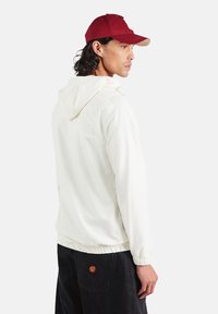 Biała kurtka z kapturem, z elastycznym ściągaczem na dole i mankietach, w połączeniu z bordową czapką z jasnobrązowym daszkiem. Czarne spodnie z małym logo.