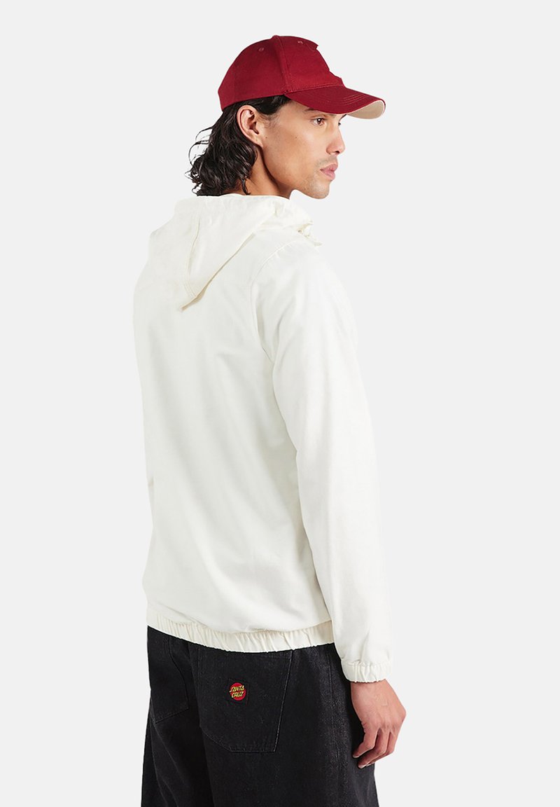 Biała kurtka z kapturem, z elastycznym ściągaczem na dole i mankietach, w połączeniu z bordową czapką z jasnobrązowym daszkiem. Czarne spodnie z małym logo.