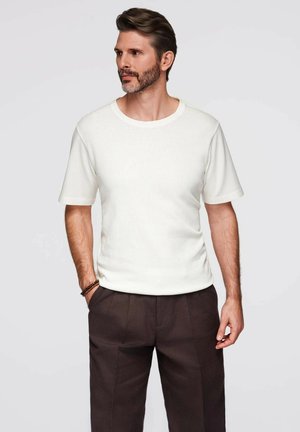 Hombre con barba, vistiendo una camisa blanca de manga corta y pantalones marrón oscuros, de pie con una mano en el bolsillo contra un fondo claro.