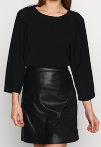 Blouse noire à manches longues avec un col rond et des plis aux épaules, associée à une jupe en cuir noir élégante avec des coutures diagonales.