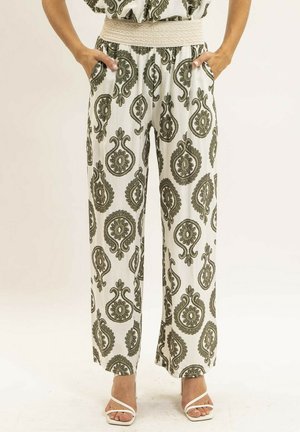 Femme portant un pantalon blanc taille haute à jambes larges, orné de motifs floraux paisley vert olive et d'une ceinture élastique texturée beige, debout les mains dans les poches.