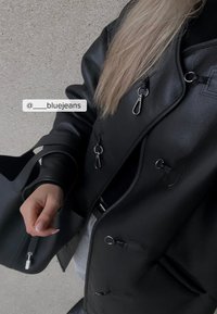 Juodas odinis švarkas su metaliniais sagtimis ir lygia tekstūra. Taip pat matoma juoda rankinė. Fone yra pilka betono danga.