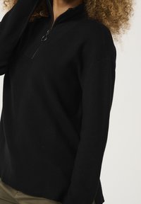 Svart tröja med kvartshals, ribbat mönster och långa ärmar. Har en avslappnad passform och veck på sidorna vid fållen.
