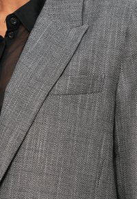 Szary blezer w jodełkę z teksturowanej tkaniny, z klapami wciętymi i pojedynczą kieszenią na piersi. Charakteryzuje się strukturalnym krojem i dopasowanym fasonem.