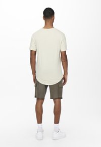 Beige T-Shirt mit kurzen Ärmeln und abgerundetem Saum, kombiniert mit olivgrünen Cargo-Shorts. Weiße Sneaker und weiße Knöchelsocken runden das Outfit ab.