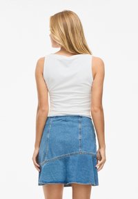 Woman with long blonde hair wearing a sleeveless white top and a blue denim skirt, standing facing away against a plain white background.