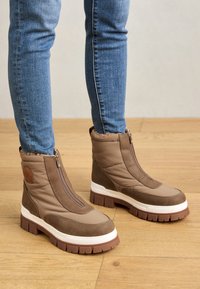 Botas de tobillo aisladas marrones con un acabado suave, cremallera vertical, suela texturada y un forro esponjoso en el cuello, combinadas con jeans azules.