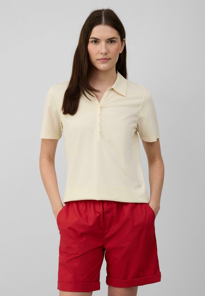 Polo shirt jaune clair, à manches courtes, avec un col boutonné. Accompagné d'un short rouge avec poches latérales et ourlets retroussés.