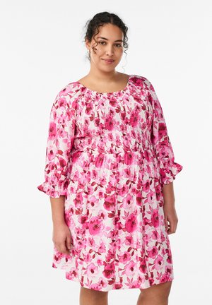 Žena s kučeravými vlasmi, ktorá nosí perlové náušnice a ružové kvetinové šaty s nahusteným živôtikom a 3/4 rukávmi s volánikmi, stojaca na bielom pozadí.