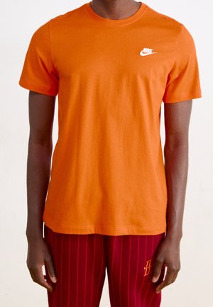 Mężczyzna w gładkiej pomarańczowej koszulce Nike i czerwonych spodniach z cienkimi pionowymi paskami oraz małym żółtym logo na udzie.