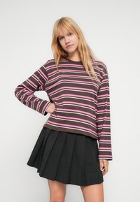 Νέα γυναίκα με μακριά ξανθά μαλλιά που φοράει μια μακρυμάνικη μπλούζα με καφέ, ροζ και λευκές ρίγες και μια μαύρη πλισέ φούστα, πόζαρε με το ένα χέρι στη μέση.