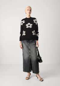 Maglione nero con motivi floreali bianchi, abbinato a una gonna maxi in denim e sandali neri. Gli accessori includono una piccola borsa nera.