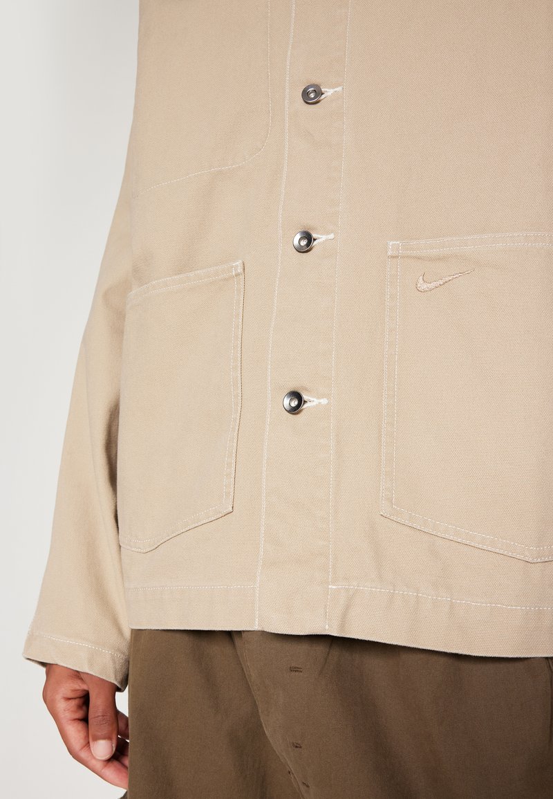Camisa beige con botones en la parte delantera, dos bolsos grandes y un sutil logo de Nike en el bolsillo derecho, combinada con pantalones marrones.