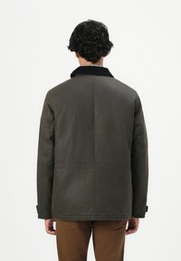 Uomo con capelli ricci corti che indossa una giacca verde scuro con colletto nero e pantaloni marroni, in piedi di spalle contro uno sfondo bianco semplice.