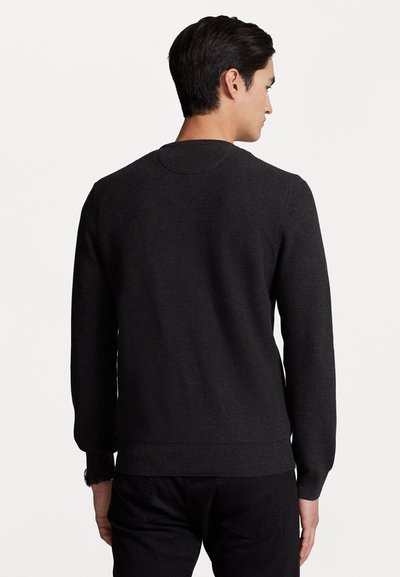 Άντρας φορεί ένα απλό μαύρο φούτερ με μακριά μανίκια και μαύρο παντελόνι, εμφανιζόμενος από πίσω σε έναν απλό λευκό φόντο.