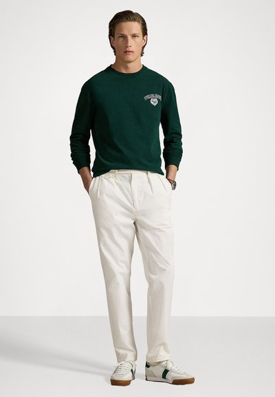 Groene sweatshirt met lange mouwen en een logo, crèmekleurige geplooide broek en witte sneakers met groene accenten. Eenvoudig ontwerp en casual stijl.