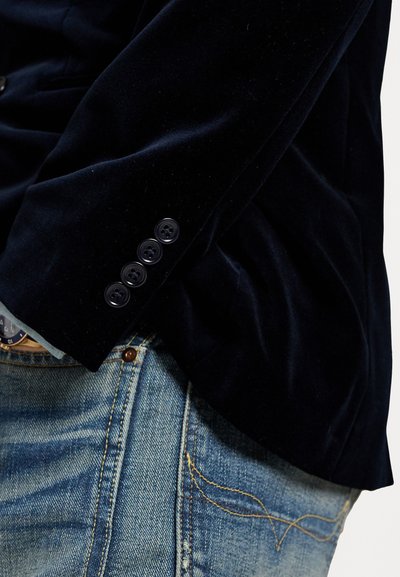 Námořnický sametový blazer s hladkou texturou, vybavený funkčními černými knoflíky na manžetách. V kombinaci s světle modrými denimovými džíny.