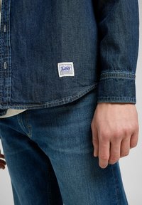 Camisa de mezclilla en azul oscuro con cuello clásico, cierre de botones y una etiqueta de trabajo blanca de Lee en la parte inferior izquierda. La textura parece suave.