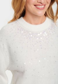 Maglione bianco con scollo rotondo, caratterizzato da un grappolo di paillettes iridescenti sulla parte superiore, che crea un effetto testurizzato.