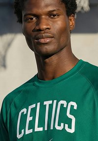 Uomo che indossa una maglia sportiva verde dei Celtics, rivolto verso l'obiettivo con espressione neutra, sfondo esterno sfocato.