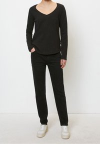 Person wearing a black long-sleeve V-neck top, black pants, and white sneakers, standing with one hand in pocket against white wall.