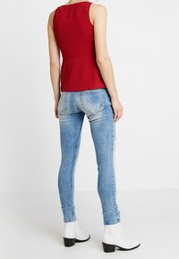 Haut sans manches rouge avec un détail froncé dans le dos, associé à un jean en denim bleu clair ajusté. Des bottes blanches à la cheville complètent le look.