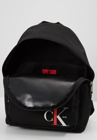 Czarny plecak materiałowy z zamkiem błyskawicznym, posiadający błyszczący czarny przedni kieszeń oraz czerwone logo. Gładka tekstura, prosty design.