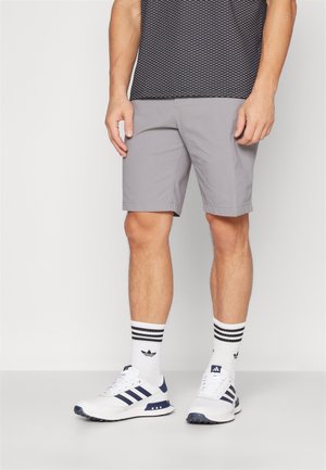 Graue Shorts mit glatter Textur, kombiniert mit einem schwarz gemusterten Hemd, weißen Sneakers mit marinefarbenen Akzenten und schwarz-weiß gestreiften Socken.