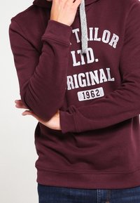 Bordeauxrode hoodie met een grijs trekkoord, voorzien van een gedrukt logo en de tekst "Tailor Ltd. Original 1962" op de borst. Zachte stoftextuur.