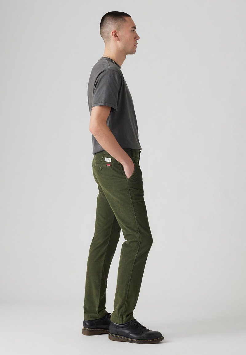 Oliwkowe bawełniane spodnie o tapered fit, z przednimi kieszeniami i tylną naszywką, zestawione z szarym t-shirtem z okrągłym dekoltem i czarnymi butami.