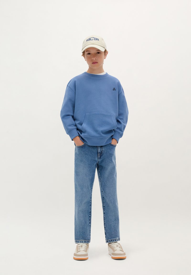 Felpa blu con tasca frontale, abbinata a jeans azzurri. Cappellino crema con testo blu e scarpe beige con dettagli bianchi.