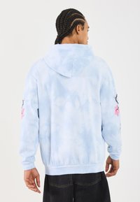 Sweatshirt azul claro em tie-dye com um corte descontraído, apresentando bordados florais em rosa na manga esquerda e capuz com cordão.
