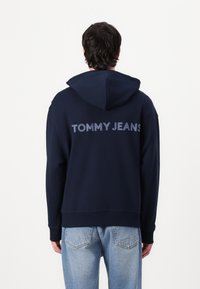 Mørkeblå hættetrøje med kengurulomme, med "TOMMY JEANS" logo i lysegrå på ryggen. Blandingsstof af bomuld, afslappet pasform.
