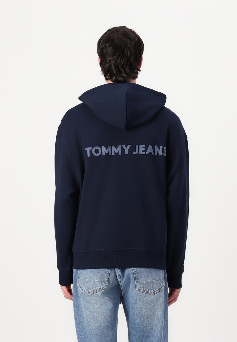 Sweat à capuche bleu marine avec poche kangourou, arborant le logo "TOMMY JEANS" en gris clair au dos. Tissu en mélange de coton, coupe décontractée.