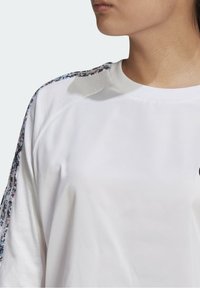 Vit sweatshirt med rund halsringning, med blommönstrade detaljer på axlarna. Tyget ser slätt och lätt ut.