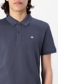 Marineblå polo t-shirt lavet af tekstureret stof, med en to-knaps knaplukning og et lille logo på venstre bryst. Korte ærmer.