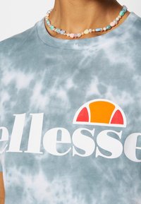 T-shirt gris tie-dye avec un logo "ellesse" blanc et un graphique de soleil coloré. Porté avec un collier de perles multicolores.