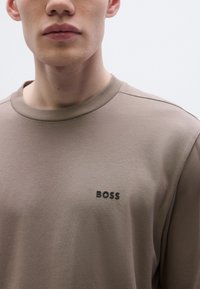 Szürke hosszú ujjú póló kerek nyakkivágással és fekete "BOSS" logóval a mellkason. Puha anyag, sima textúrával.