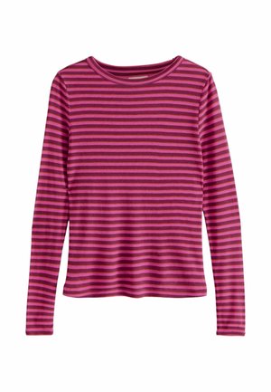 Femme portant une chemise à manches longues rayée rose et rouge foncé et un pantalon noir, avec une main dans la poche, sur un fond uni.